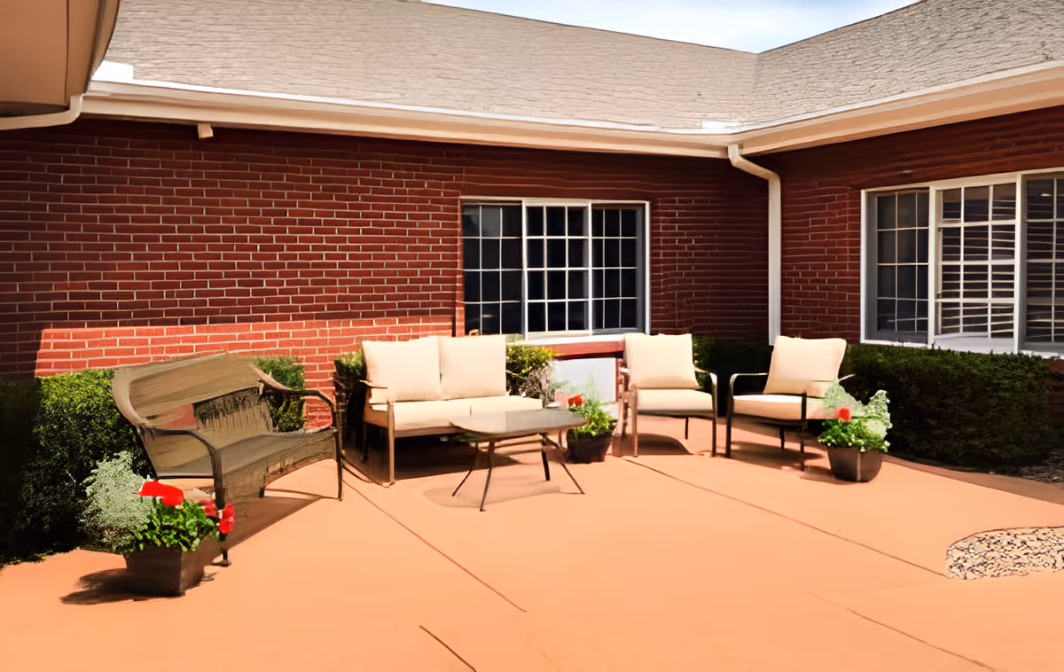 Outdoor patio area with cushioned seating including a loveseat, two armchairs, and a metal bench. There is a small coffee table in the center and potted plants with red flowers placed around the seating area. The patio is adjacent to a brick building with large windows and trimmed hedges along the walls.