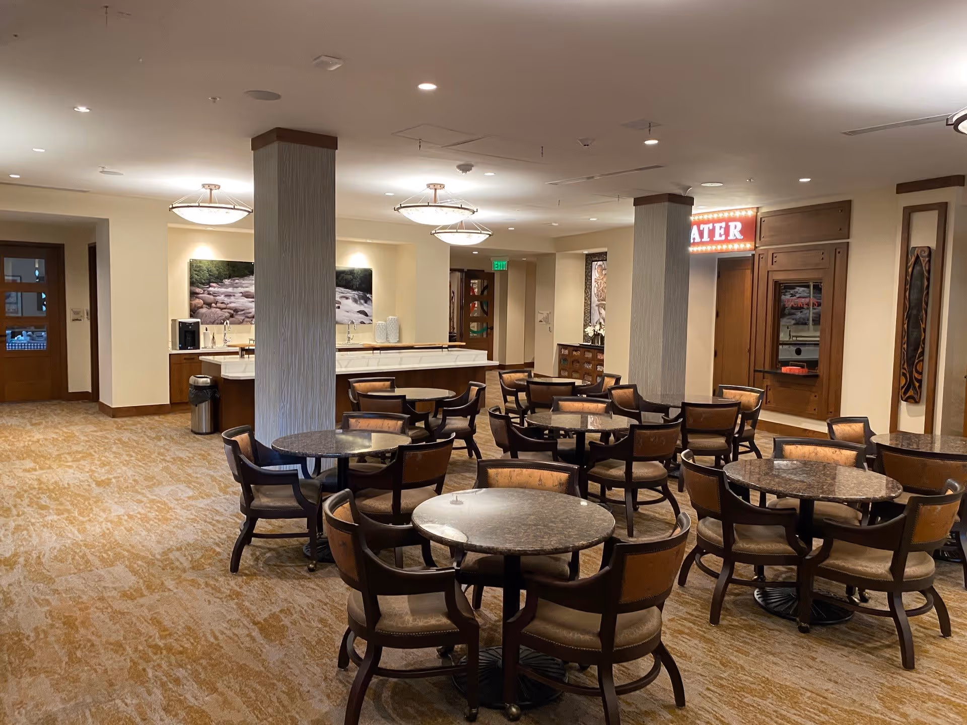 A spacious dining area with multiple round tables and cushioned chairs arranged neatly. The room has beige walls, carpeted floors, and ceiling lights. There is a counter area with a coffee machine and decorative items, and a sign with the word 'THEATER' illuminated in red lights is visible on the right side.