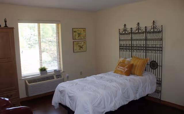 A simple bedroom with a single bed covered in a white textured comforter and two yellow pillows, one with the words 'Family Forever'. The bed has a decorative black metal headboard. There is a window with blinds partially open, a small plant on the windowsill, and two framed pictures on the wall. A wooden cabinet is visible on the left side of the room.