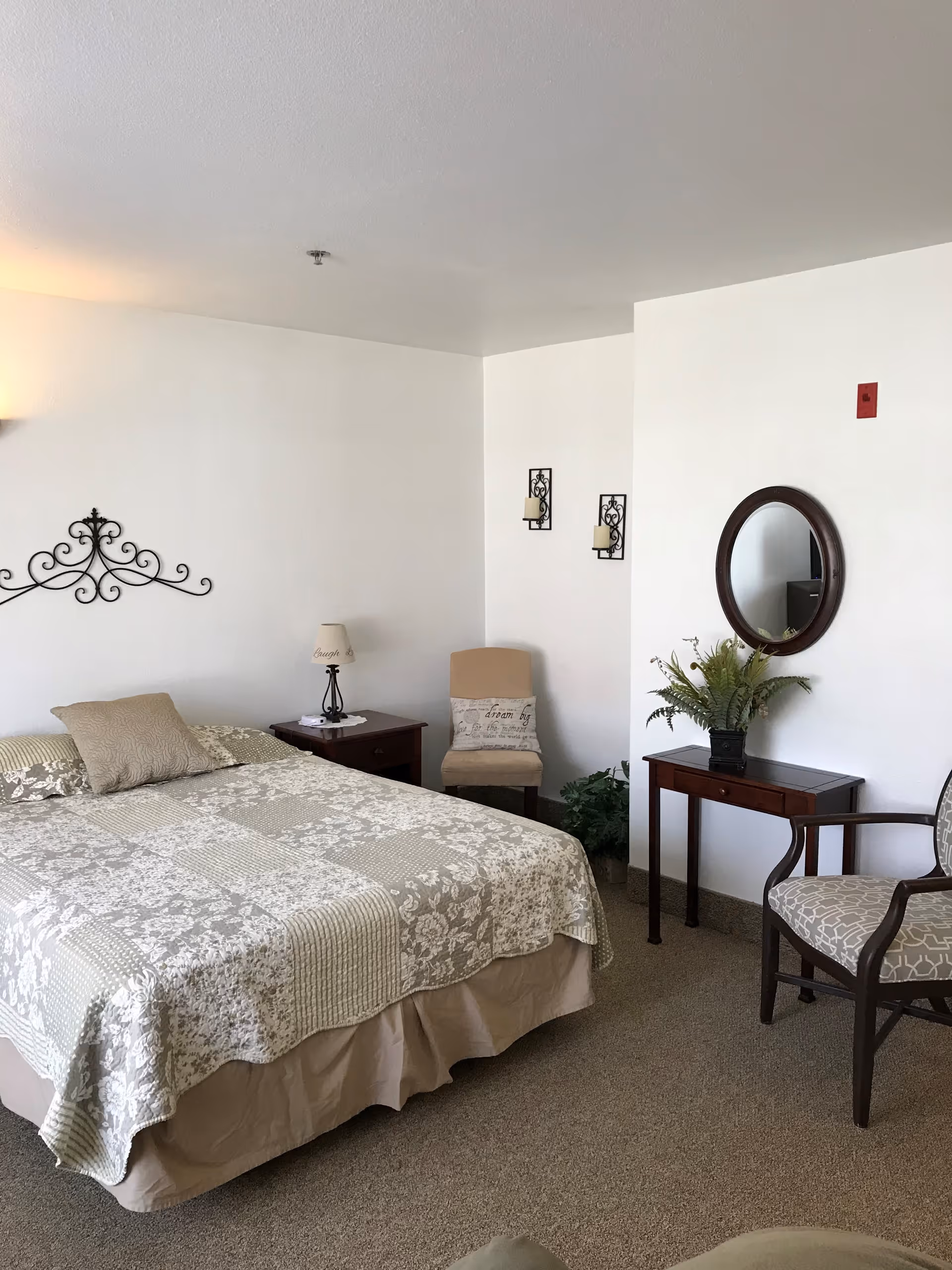 A bedroom with a bed covered in a beige and white floral quilt, a beige pillow, a wooden nightstand with a lamp, a beige chair with a decorative pillow, a small wooden table with a plant, a round mirror on the wall, and a wooden armchair with patterned upholstery.