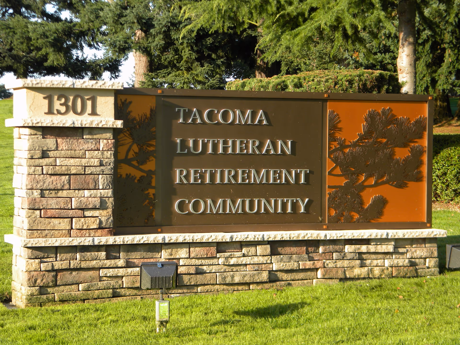 Stone and metal sign for Tacoma Lutheran Retirement Community with the address number 1301, surrounded by green grass and trees in the background.