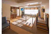 A well-lit therapy or rehabilitation room with parallel bars for walking exercises, a blue armchair, wooden cabinets, a treadmill, and a wall-mounted TV. The room has large windows with blinds allowing natural light to fill the space.