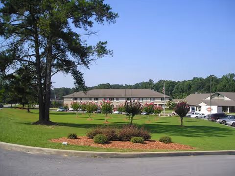 View of a senior living facility building with a large green lawn and several small trees in the foreground. The building has a beige exterior with a brown roof, and there are several cars parked near the entrance. Tall pine trees are visible on the left side, and the sky is clear and blue.