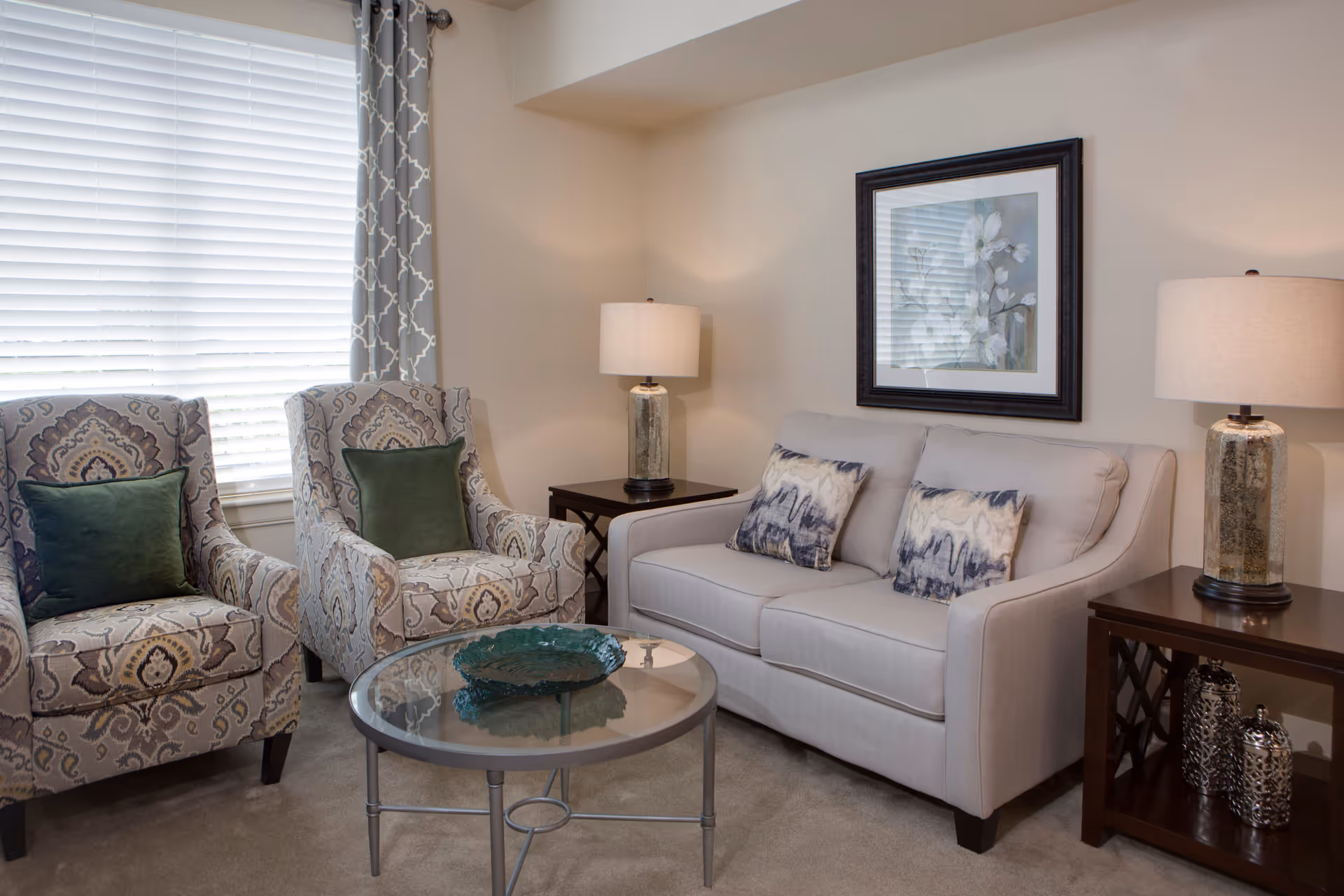 A cozy living room with a light gray sofa adorned with two patterned pillows, two patterned armchairs each with a green cushion, a round glass coffee table with a decorative bowl, two side tables with matching lamps, and a framed floral artwork on the wall.