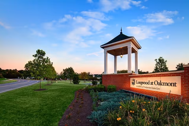 Landscaped entrance with a brick sign reading "Longwood at Oakmont", a small white gazebo, and a curved road under a colorful evening sky.