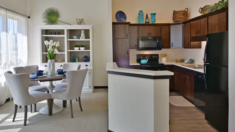 A bright and modern kitchen and dining area in a senior living facility. The kitchen features dark wood cabinets, a black refrigerator, stove, microwave, and dishwasher. The dining area has a round table set with blue dishes and white chairs, next to a white cabinet with decorative items and a large window with sheer curtains letting in natural light.