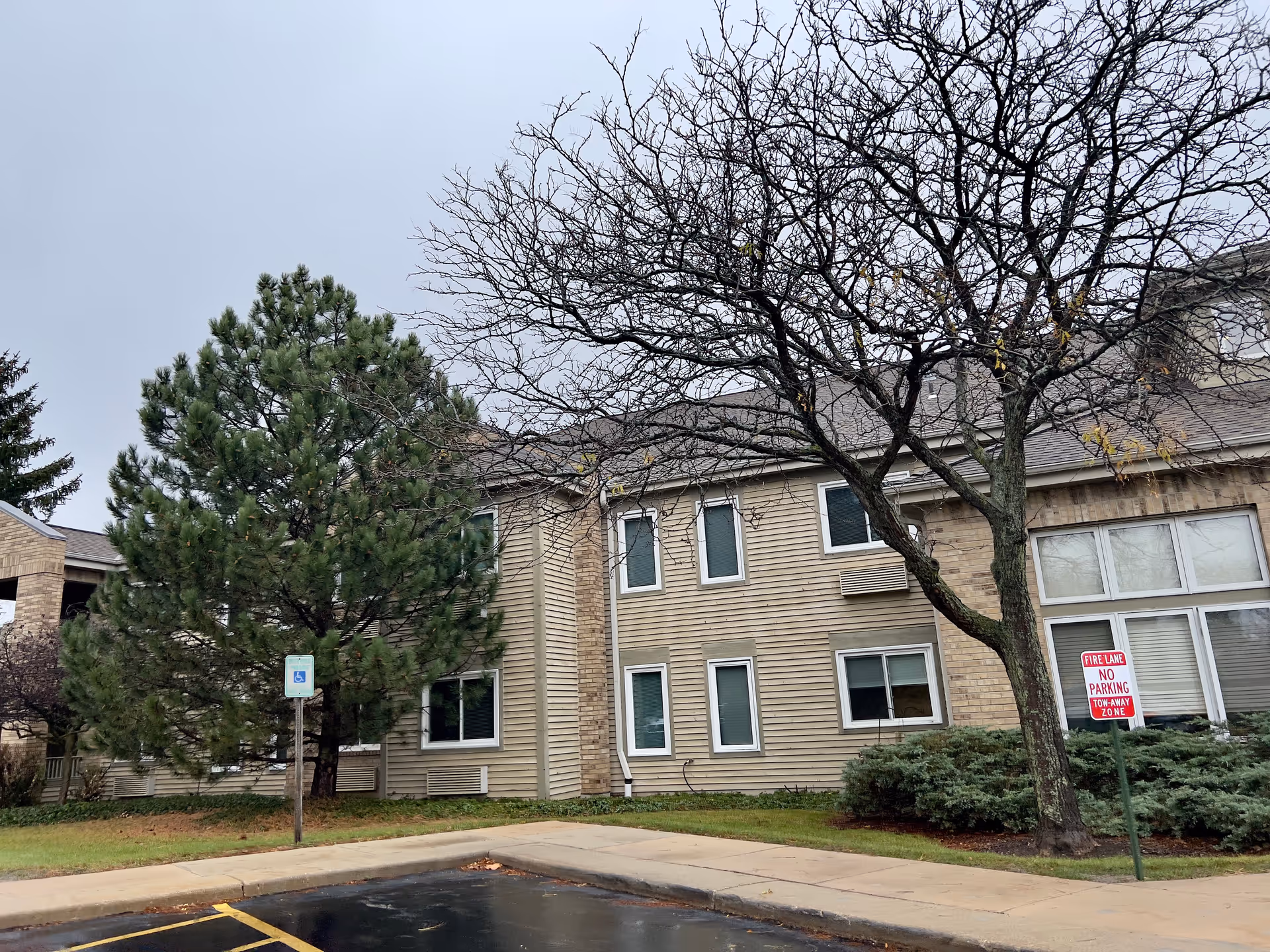 Exterior view of a two-story building with beige siding and brick accents, surrounded by trees including a large pine and a leafless tree. There is a sidewalk and a parking area with a handicapped parking sign and a fire lane no parking sign.