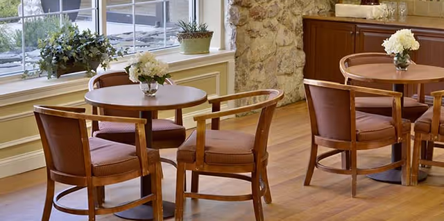 Dining area with round wooden tables and upholstered chairs, floral centerpieces, a window, and a stone accent wall.