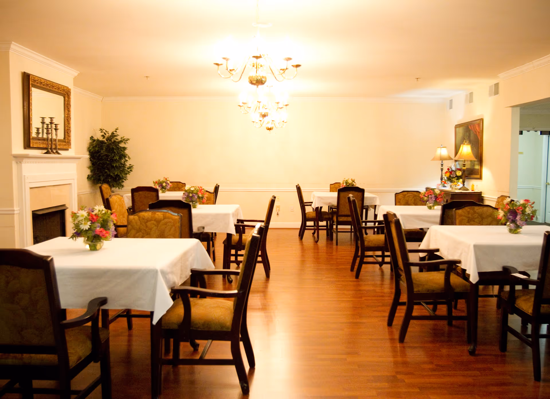 A well-lit dining room with several tables covered in white tablecloths, each adorned with a small floral centerpiece. The room features wooden chairs with cushioned seats, a hardwood floor, a fireplace with a mirror above it, a potted plant, and a side table with lamps and flowers. The walls are painted a light color, and chandeliers hang from the ceiling.