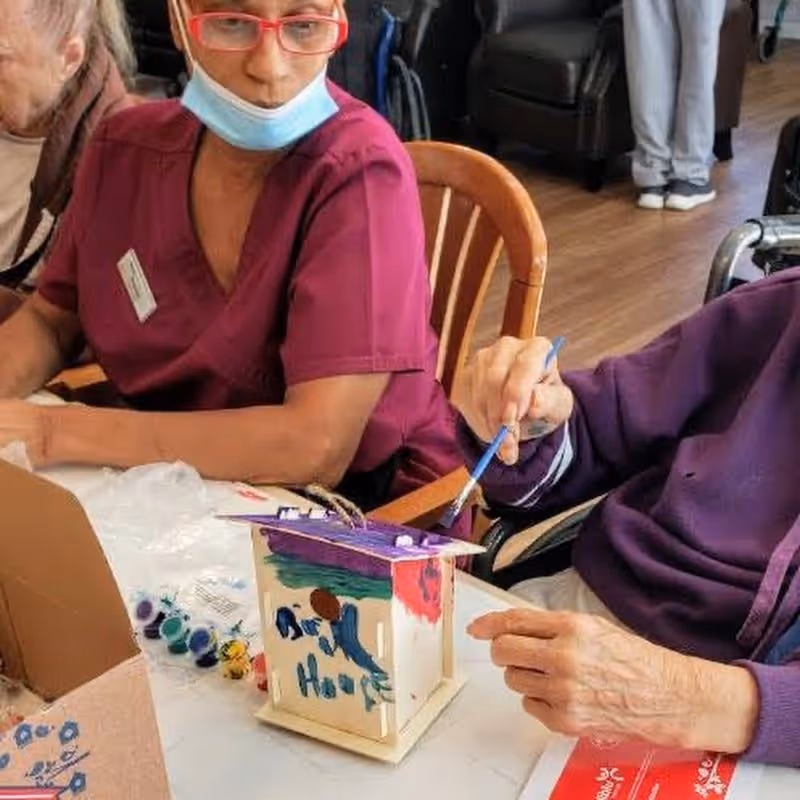 An elderly person painting a small wooden house model with a paintbrush, assisted by a caregiver wearing a maroon uniform and a face mask pulled down below her chin, seated at a table with paint supplies.