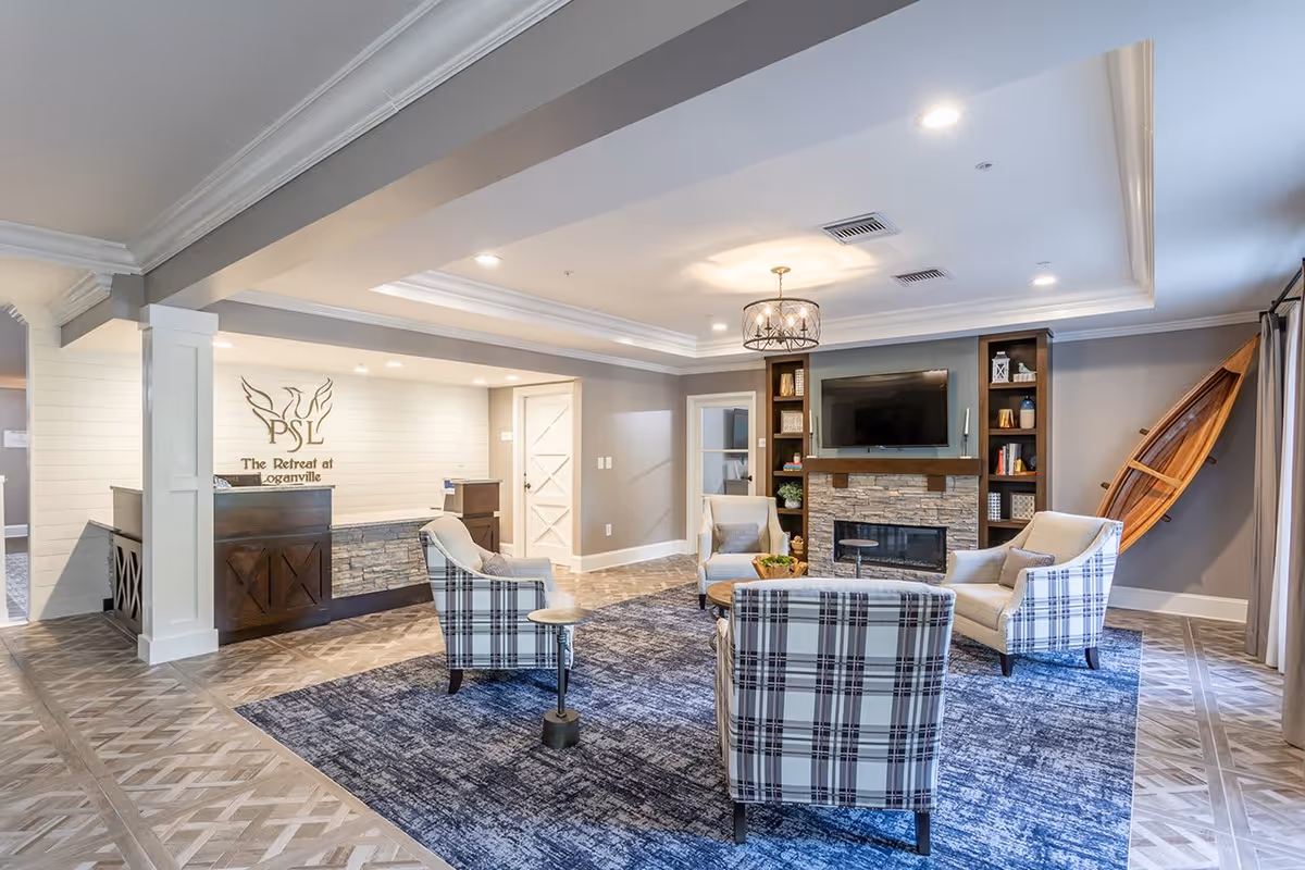 A bright and spacious living room area in The Retreat at Loganville featuring a stone fireplace with a mounted TV above it, built-in shelves with decorative items, four plaid armchairs arranged around a wooden coffee table on a blue area rug, and a wooden canoe mounted on the wall. The reception desk with the facility's logo is visible in the background.