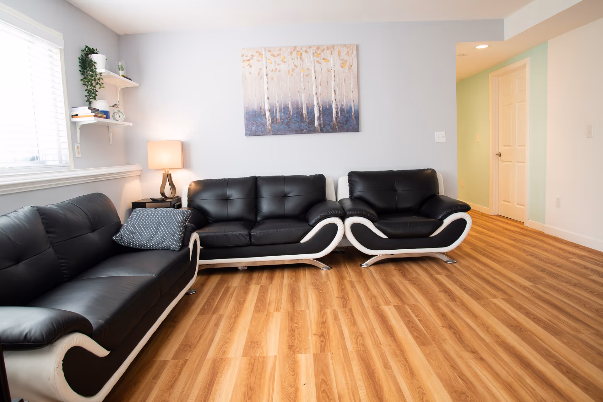 A bright living room with two black and white leather sofas, a wooden floor, a side table with a lamp, a wall painting of birch trees, and a window with white blinds. There are also two white shelves with books and plants on the left wall.