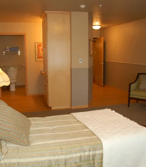 A cozy bedroom in a senior living facility with a bed covered in striped bedding and a white blanket. To the left, there is an open door leading to a bathroom with a toilet and towel rack visible. The room has wooden flooring, a chair with green upholstery, and a cabinet dividing the bedroom from the hallway.
