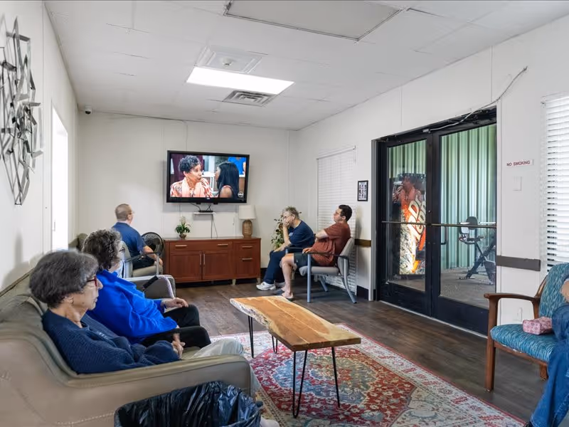 A group of elderly people sitting in a living room watching a television mounted on the wall. The room has a wooden coffee table on a patterned rug, a wooden cabinet under the TV, and large glass doors leading to an outdoor area with a vending machine visible outside. The walls are white, and the floor is wooden.