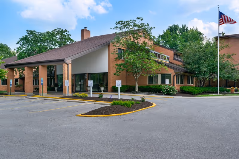 Exterior view of a brick senior living facility with trees, parking, and a flag.