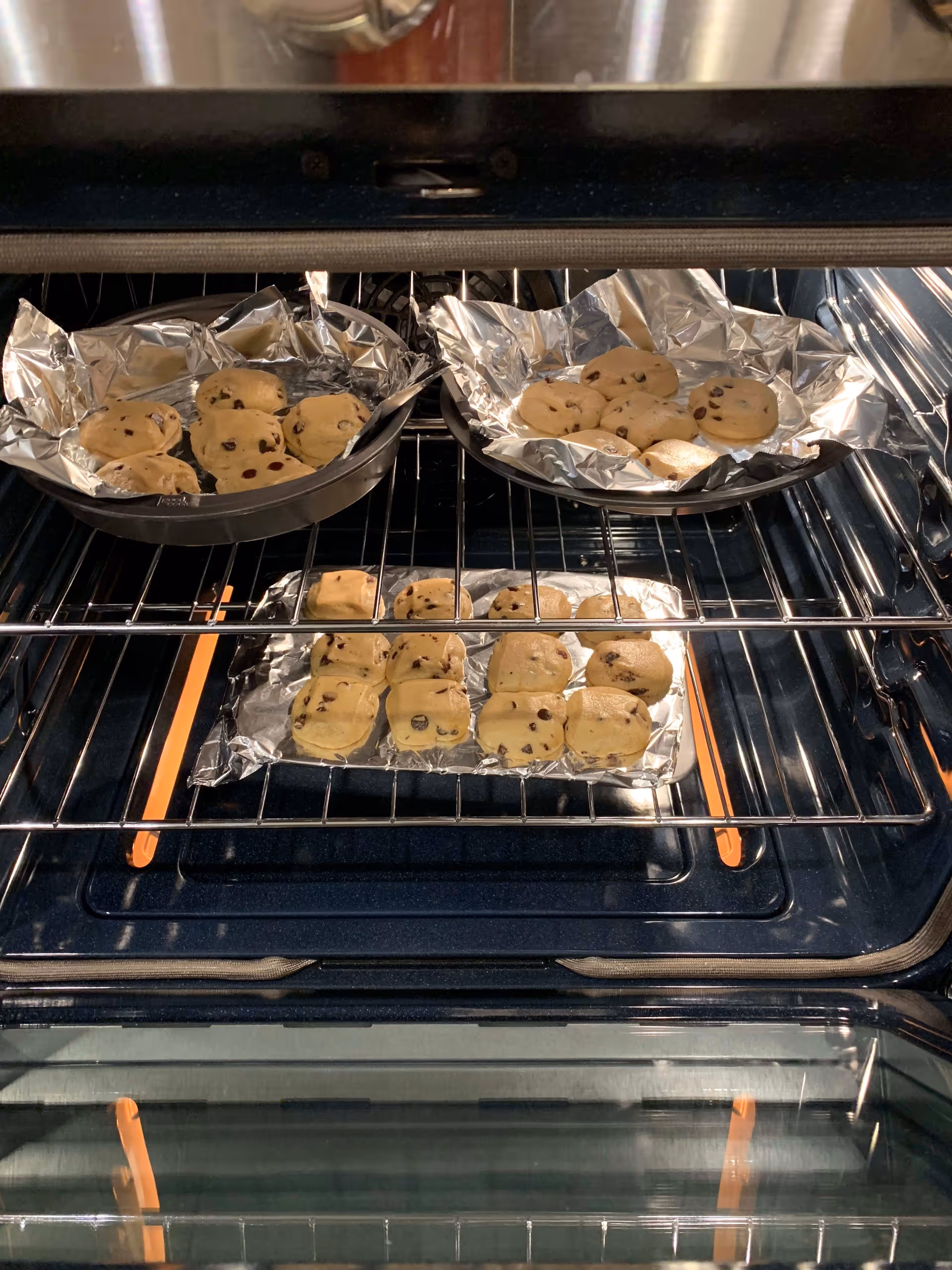 Inside view of an oven with three trays of chocolate chip cookie dough placed on aluminum foil, baking on the oven racks.