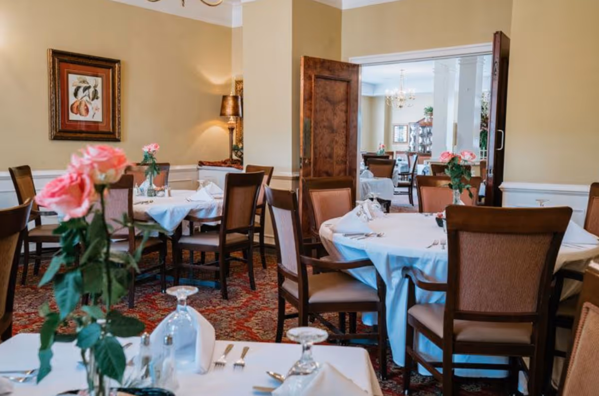 A formal dining room with round tables covered in white tablecloths, wooden chairs, and vases of pink roses.