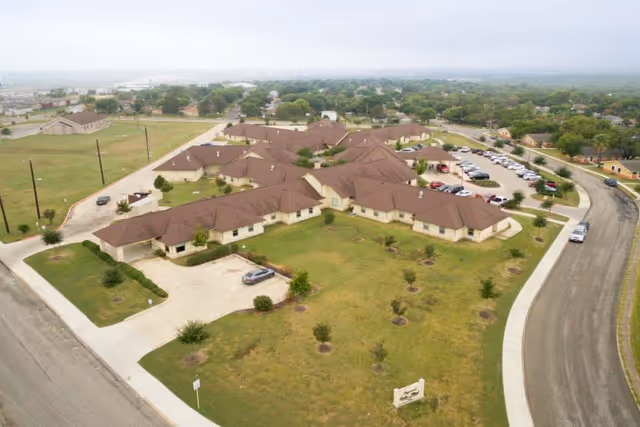 Aerial view of Legend Oaks Healthcare and Rehabilitation - West San Antonio, showing a large single-story building with multiple brown roofs surrounded by green lawns, a parking lot with cars, and a curved road.
