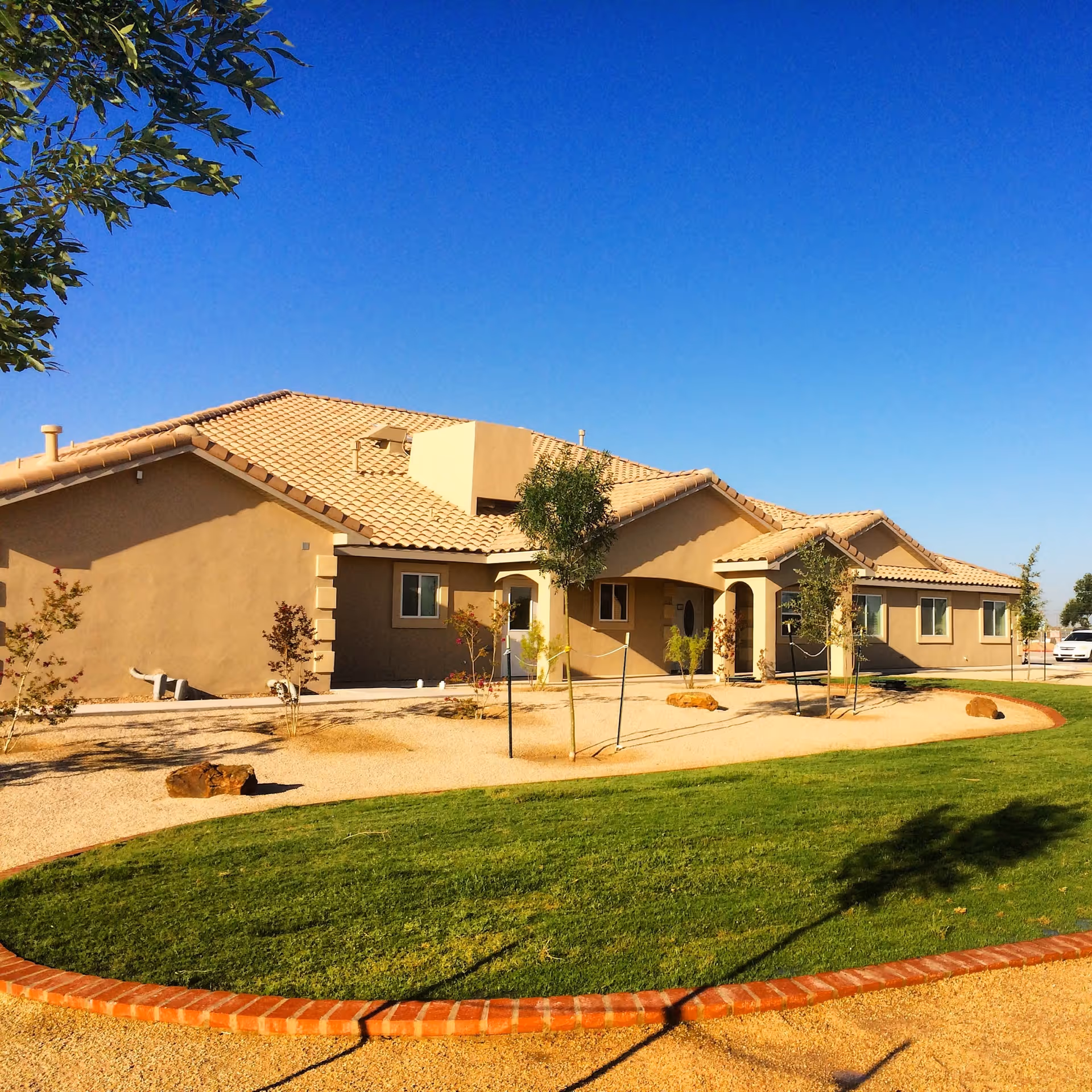 Exterior view of a single-story senior living facility building with beige stucco walls and a tiled roof, surrounded by a landscaped yard with green grass, small trees, and a clear blue sky.