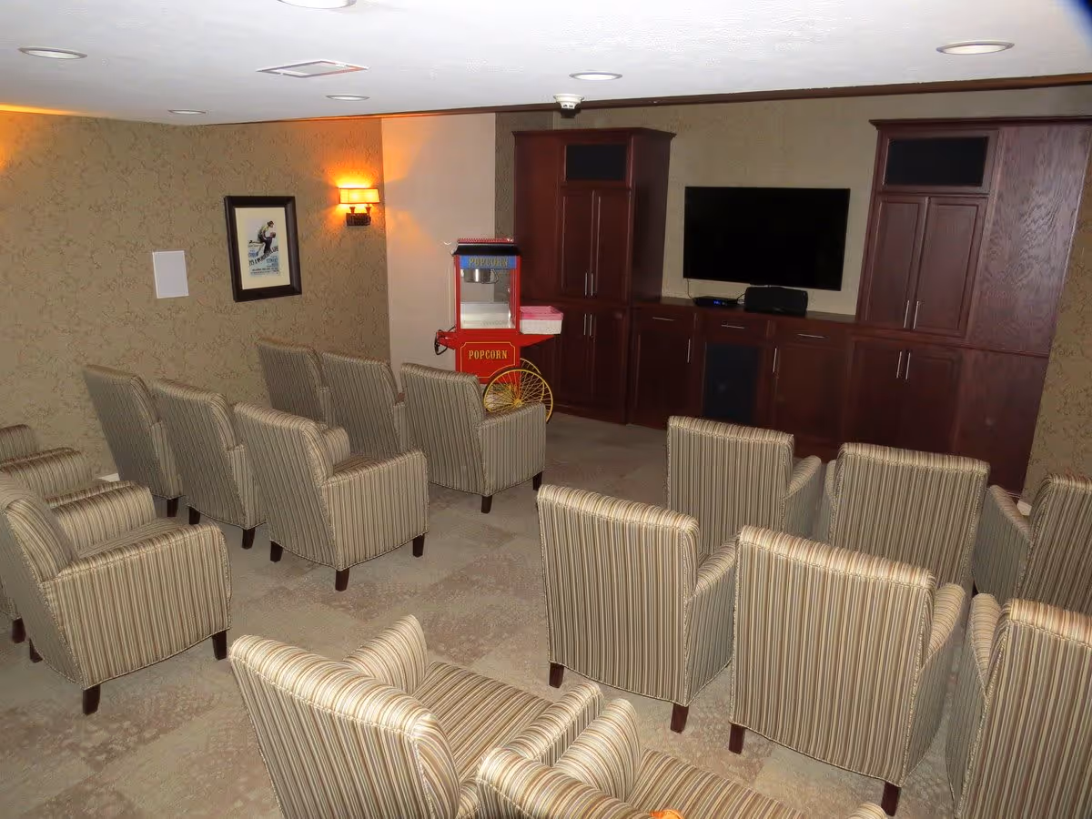 A small theater room with two rows of striped armchairs facing a flat-screen TV mounted on a wall with dark wooden cabinets. A red popcorn machine is positioned near the corner, and a framed picture and wall sconce light are visible on the left wall.