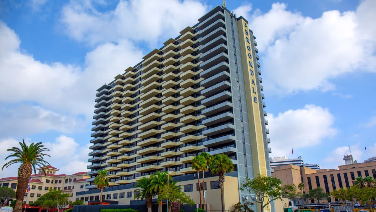 Tall multi-story building with balconies on each floor under a partly cloudy blue sky. The building has the word 'BROOKDALE' displayed vertically on one side. Palm trees and other greenery are visible in front of the building, along with other smaller buildings in the background.