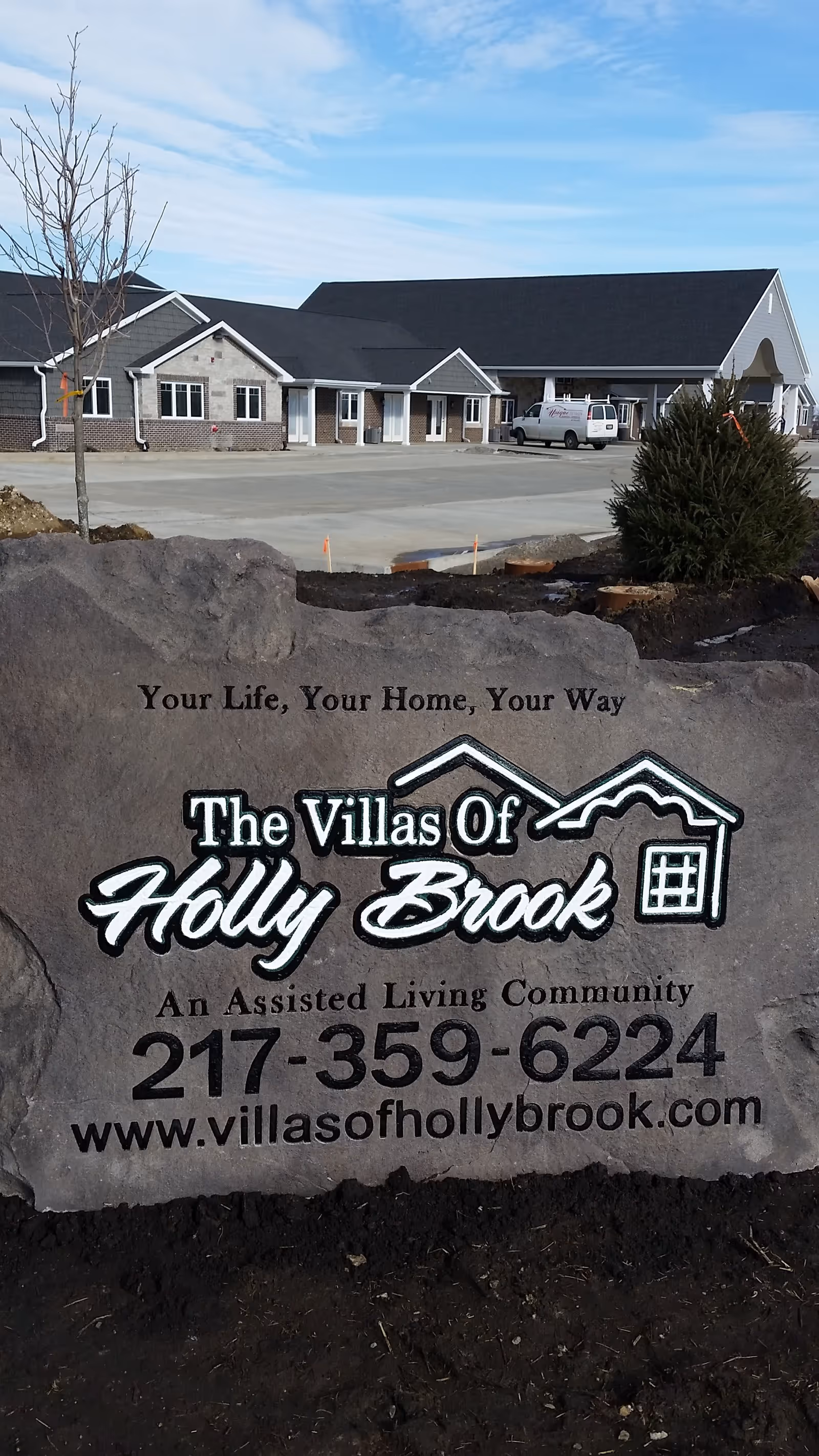A large stone sign in front of a building with a parking lot. The sign reads 'Your Life, Your Home, Your Way. The Villas Of Holly Brook. An Assisted Living Community. 217-359-6224. www.villasofhollybrook.com'. The building in the background appears to be a single-story facility with multiple windows and a covered entrance.