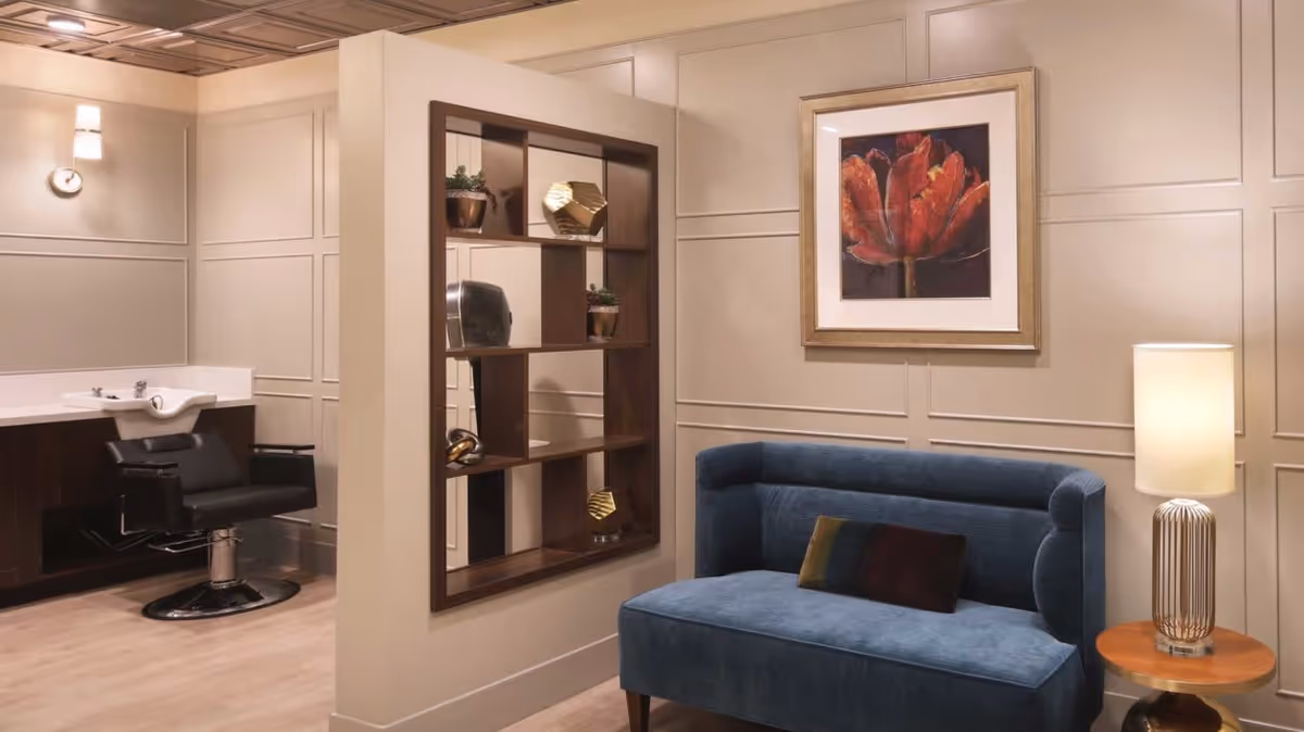 A stylish interior space featuring a blue upholstered loveseat with a small decorative pillow, a wooden side table with a modern lamp, a wall-mounted shelving unit with decorative items, and a black salon chair next to a white sink basin. The walls are paneled and painted in a neutral tone, with a framed artwork of a red flower hanging above the loveseat.