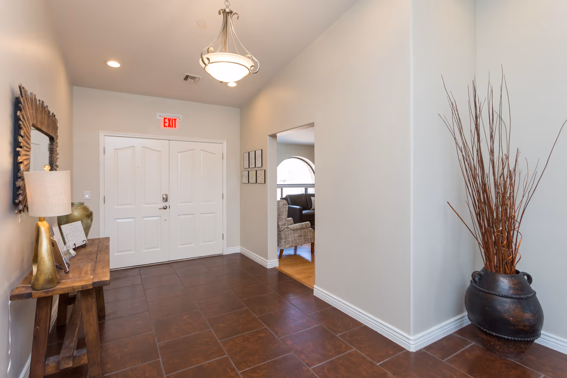 Interior hallway of Desert Gardens Assisted Living and Memory Care with brown tiled floor, white double doors with an exit sign above, a wooden console table with a lamp and decorative items on the left wall, a large decorative mirror above the table, and a large vase with tall dried branches on the right side. An open doorway leads to a room with chairs and a window.