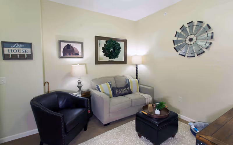 A cozy living room with a beige loveseat adorned with three pillows, one of which says 'FAMILY'. Next to the loveseat is a black leather armchair and a small side table with a lamp. On the wall above the loveseat are three decorative pieces: a framed picture of a barn, a mirror with a green wreath, and a large metal windmill wall art. A black ottoman with a tray holding decorative items sits on a patterned rug in front of the loveseat. The walls are painted light beige.