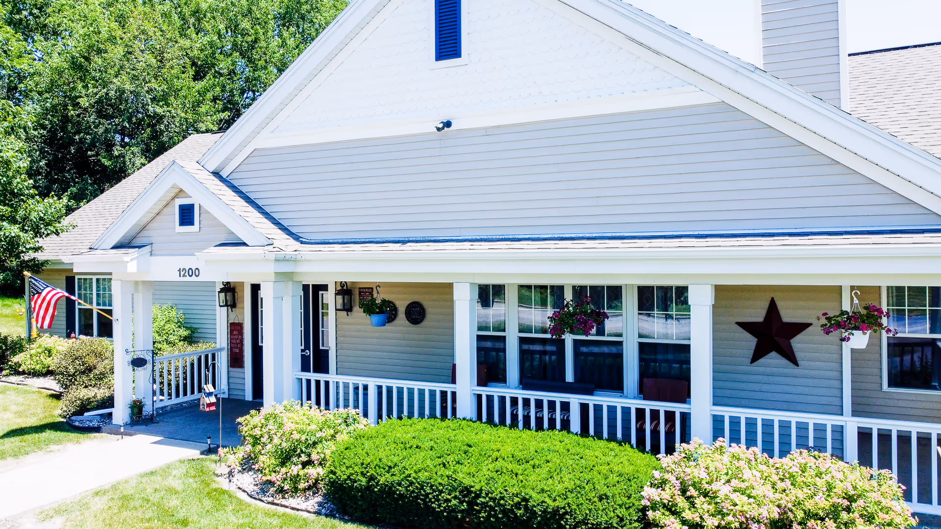 Front exterior of a light-gray assisted living building with a covered porch, white railing, hanging flower baskets, an American flag, and shrubs.
