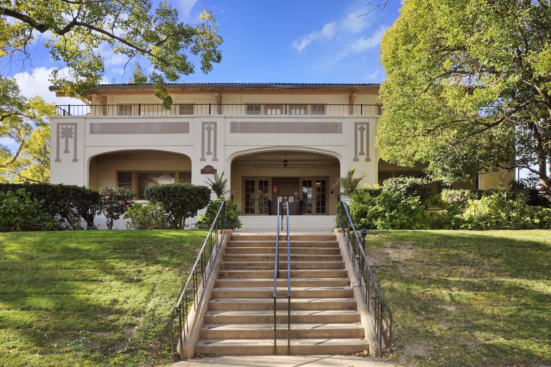 Front exterior view of a two-story assisted living facility building with a staircase leading up to the entrance. The building is surrounded by green bushes and trees under a blue sky with some clouds.