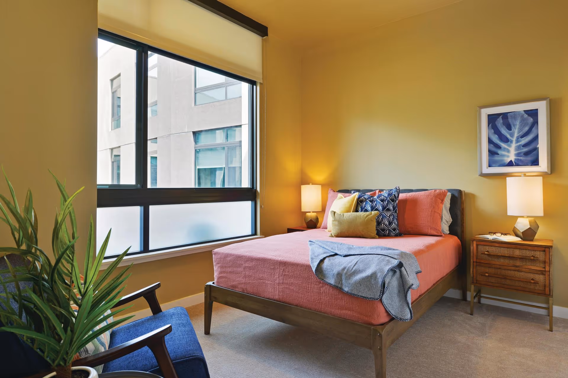 Sunlit furnished bedroom with a bed, bedside tables and lamps, a window, chair, and potted plant.