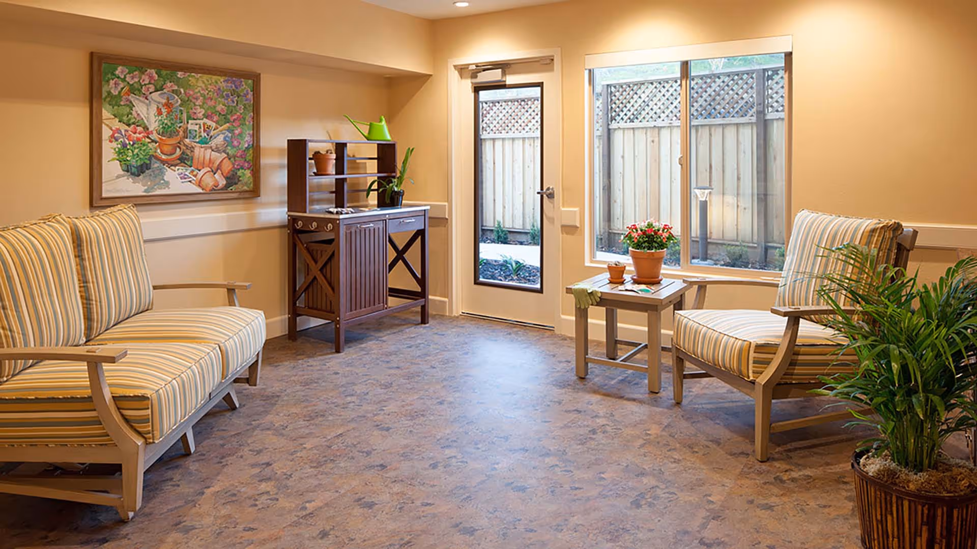 A cozy sitting area with two striped cushioned wooden chairs and a small wooden table between them holding a potted plant and gardening gloves. There is a wooden cabinet with shelves and a green watering can on top against the wall. A large window and a glass door provide a view of a fenced outdoor garden area. A colorful floral painting hangs on the wall above one chair, and a potted plant is placed on the floor near the other chair.