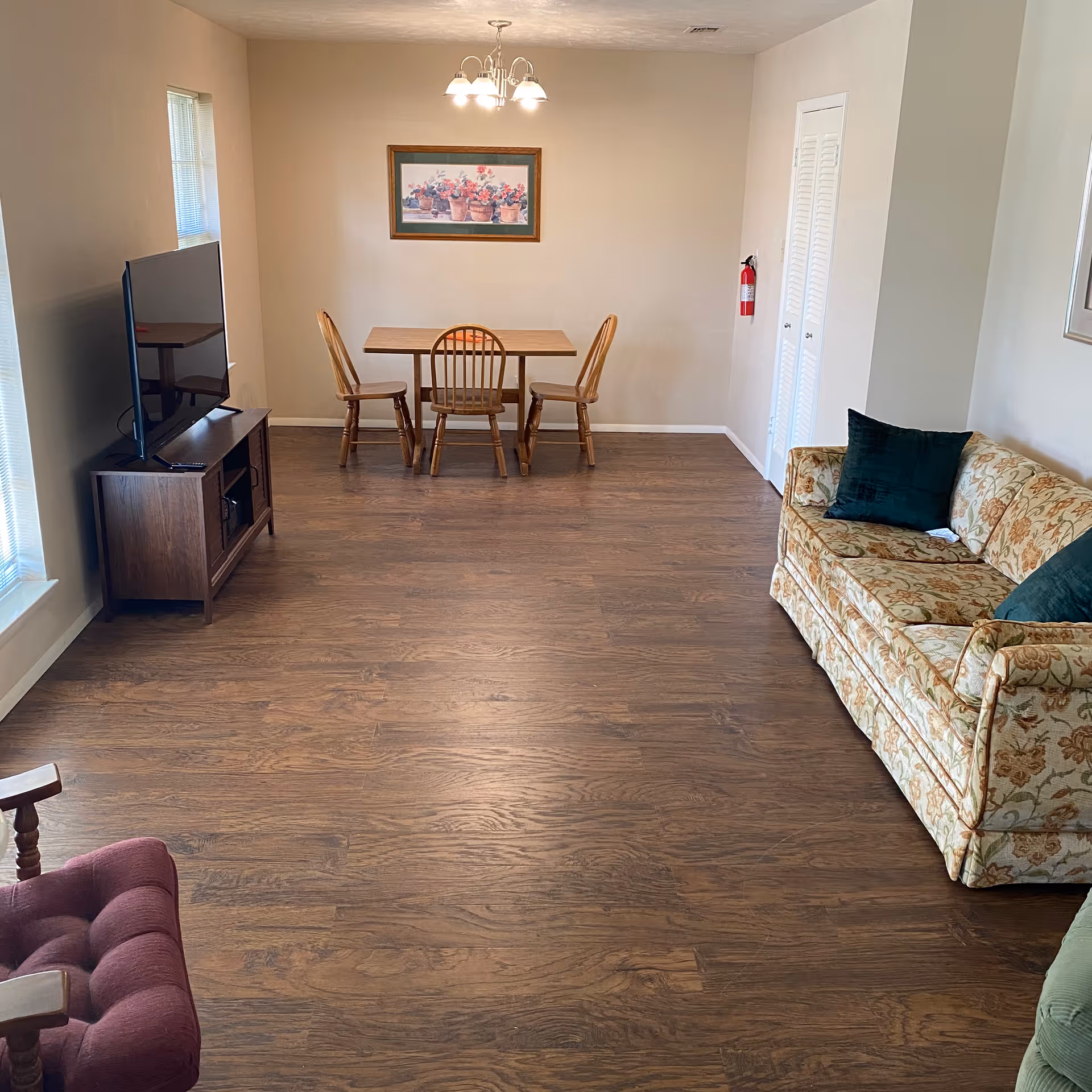 A senior living facility common area with wooden flooring, a floral patterned sofa with dark green cushions on the right, a wooden dining table with three chairs against the far wall, a flat-screen TV on a wooden stand to the left, and a framed picture of potted flowers hanging on the wall above the dining table. A fire extinguisher is mounted on the wall near a white closet door.