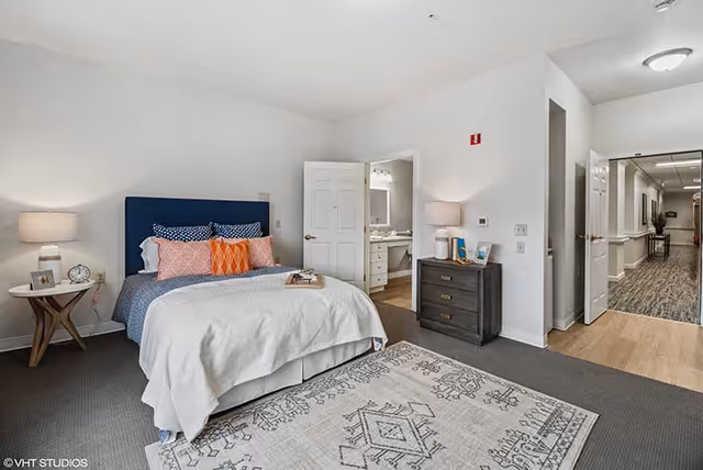 Furnished bedroom with a bed, accent pillows, nightstands, a dresser, an open bathroom door, and a hallway visible.