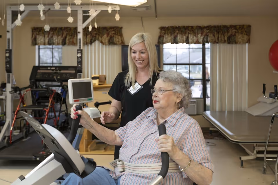 An elderly woman using a stationary exercise bike with a safety belt, assisted by a smiling female healthcare worker in a rehabilitation or physical therapy room with exercise equipment and a treatment table in the background.