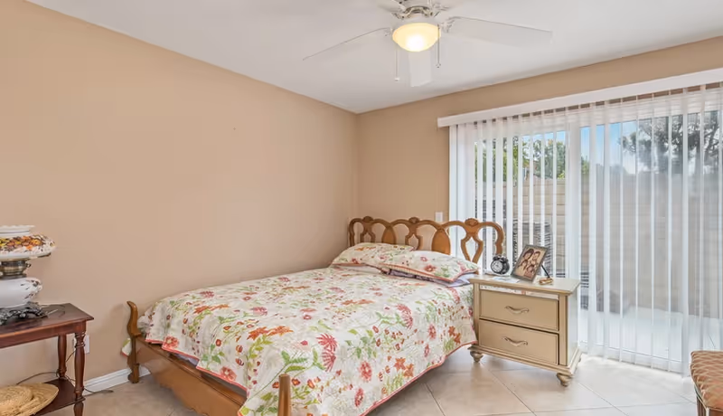 A bedroom with a wooden bed frame and floral bedspread, a nightstand with a clock and framed photo, a table lamp on a small wooden table, beige walls, tiled floor, ceiling fan, and vertical blinds covering a sliding glass door.