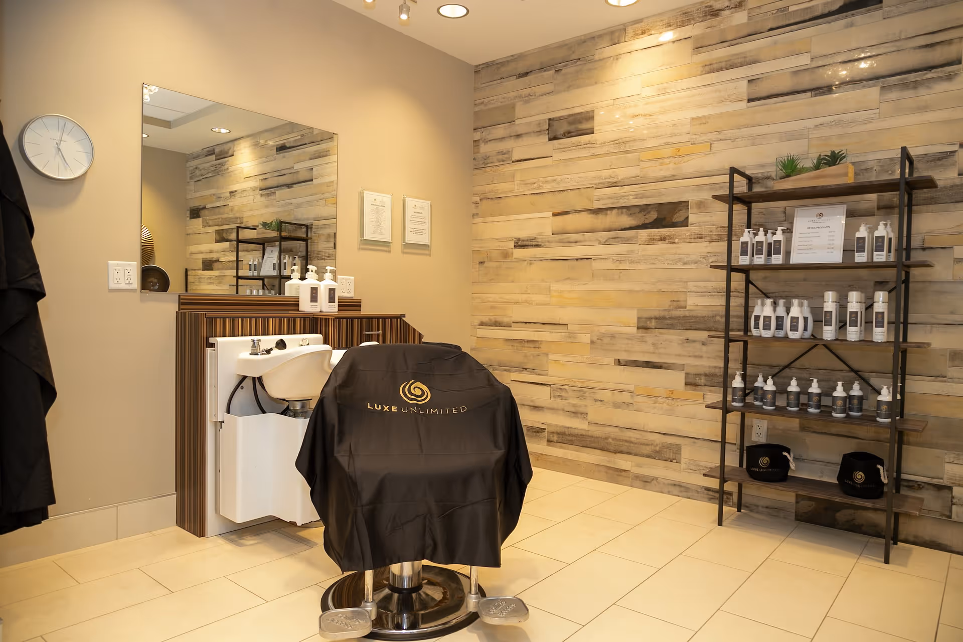 Interior view of a hair salon area with a styling chair covered by a black cloth with the logo 'Luxe Unlimited'. There is a white sink attached to a wooden counter with hair care products on top. A large mirror is mounted on the wall above the counter. On the right side, there is a black metal shelf holding various bottles and containers of hair care products. The wall behind the shelf has a rustic wooden plank design, and the floor is tiled in a light color.
