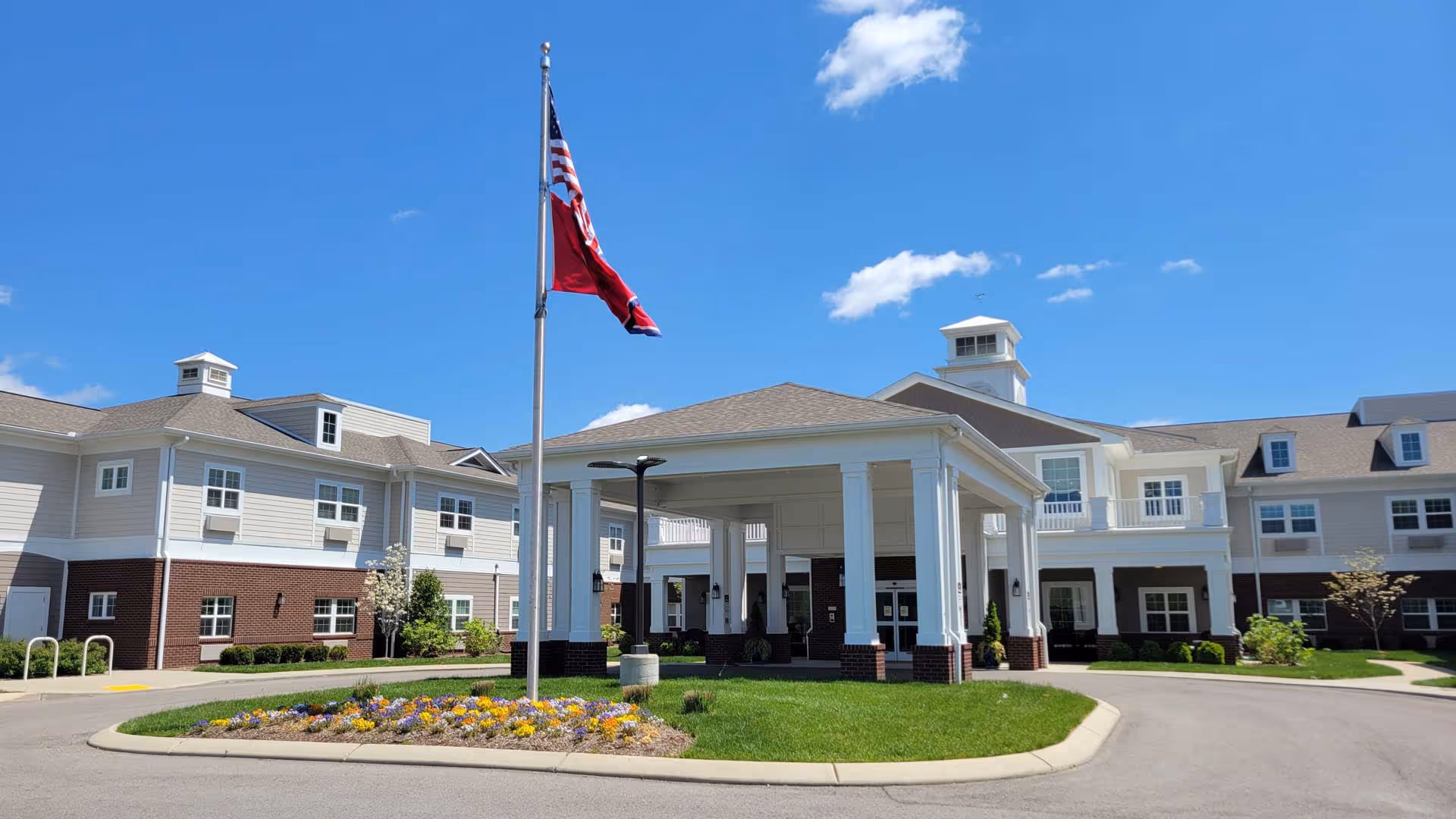 Front exterior view of The Goldton at Spring Hill facility with a covered entrance, a flagpole with two flags, a flower bed, and a clear blue sky.