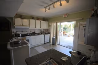 Interior view of a kitchen with white cabinets, a stove, microwave, and refrigerator. There is a countertop with some items on it, and a sliding glass door leading to an outdoor patio area with potted plants and seating.
