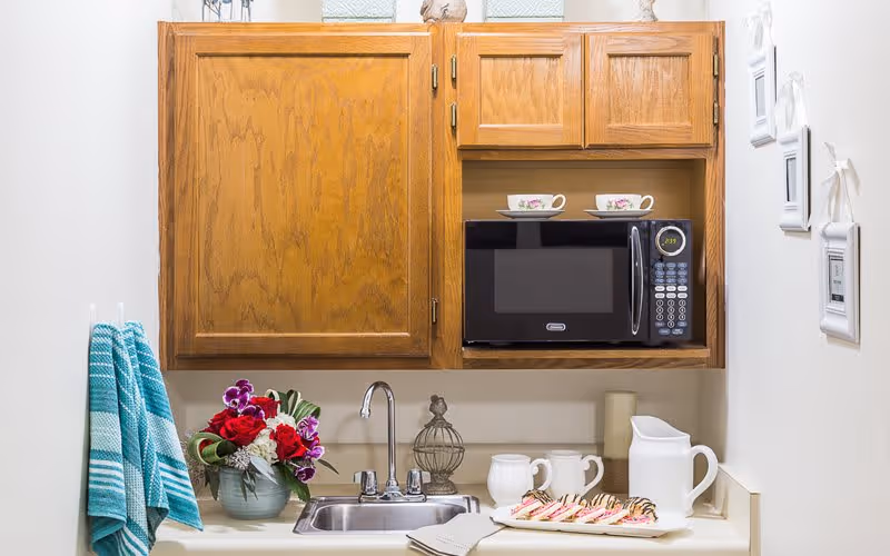 A small kitchen area with wooden cabinets, a microwave, a stainless steel sink, a bouquet of flowers in a vase, two striped blue and white towels hanging on the left, and a plate of decorated cookies with a white pitcher and two mugs on the countertop.