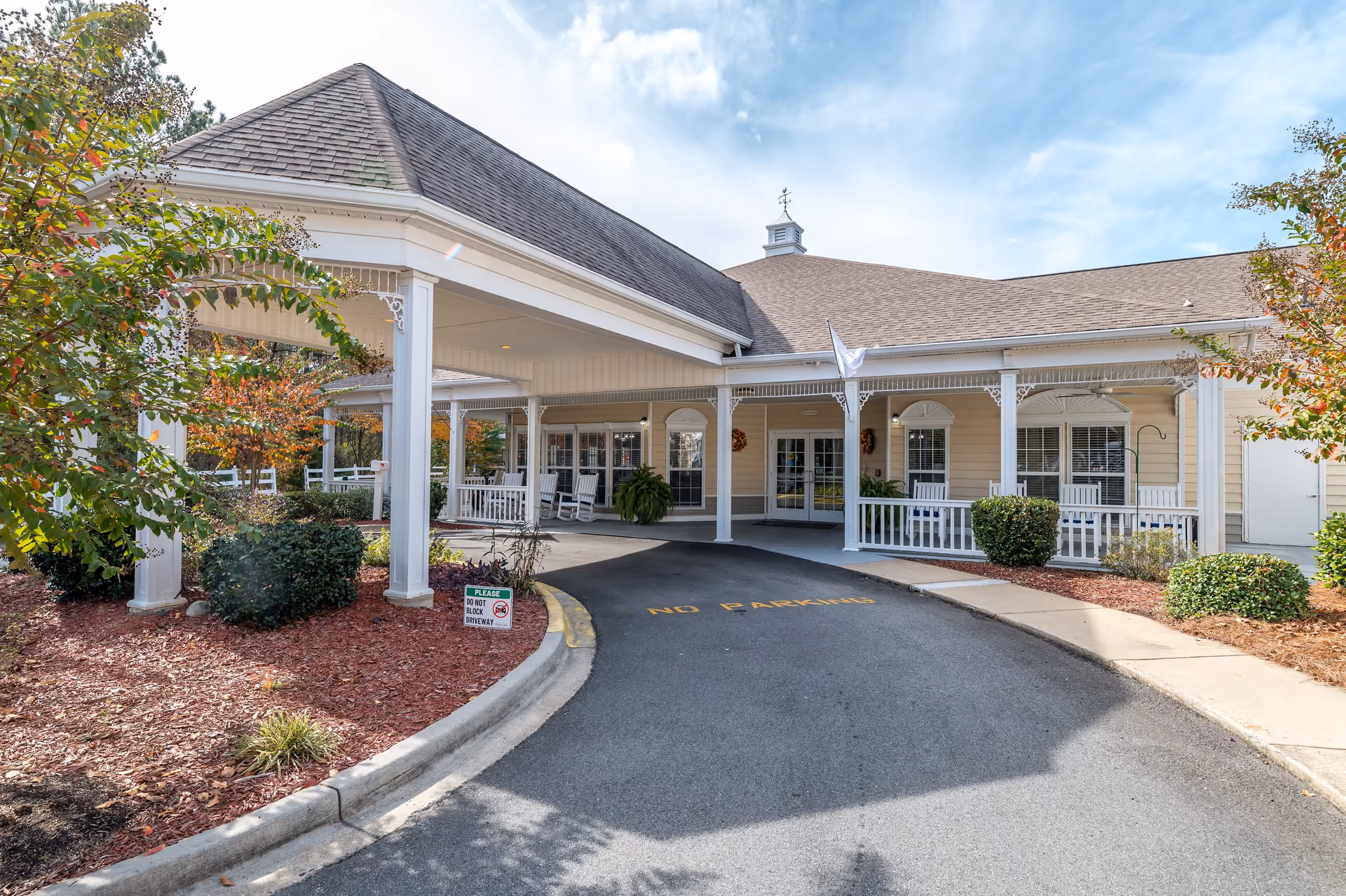 Entrance of a senior living facility with a covered driveway, white pillars, rocking chairs on the porch, and landscaped bushes and trees with autumn foliage.