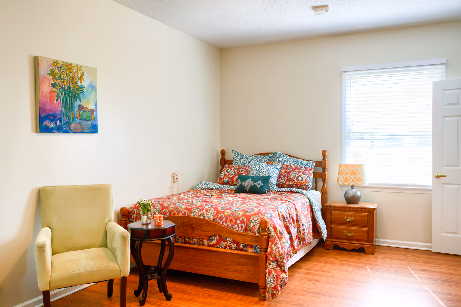 A cozy bedroom with a wooden bed featuring a colorful red and blue patterned bedspread and multiple pillows. Next to the bed is a wooden nightstand with a lamp and a window with blinds allowing natural light to fill the room. A light green upholstered armchair is positioned beside a small round wooden table with a plant and a cup. A colorful painting of flowers and fruit hangs on the wall above the chair.