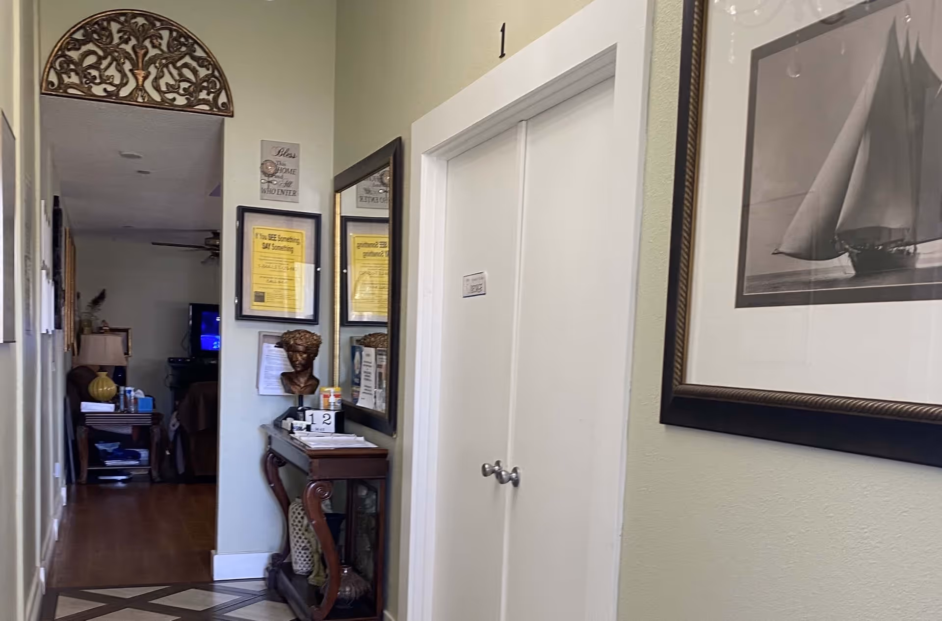 Interior hallway of a senior living facility with light green walls, a white double-door closet labeled with the number 1, a decorative wooden console table with a bust sculpture and various papers, a large mirror, framed signs on the wall, and a framed black and white picture of a sailboat. The hallway leads into a living room area with a TV and furniture visible in the background.