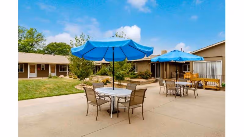 An outdoor courtyard with round patio tables, chairs and bright blue umbrellas in front of single-story buildings.