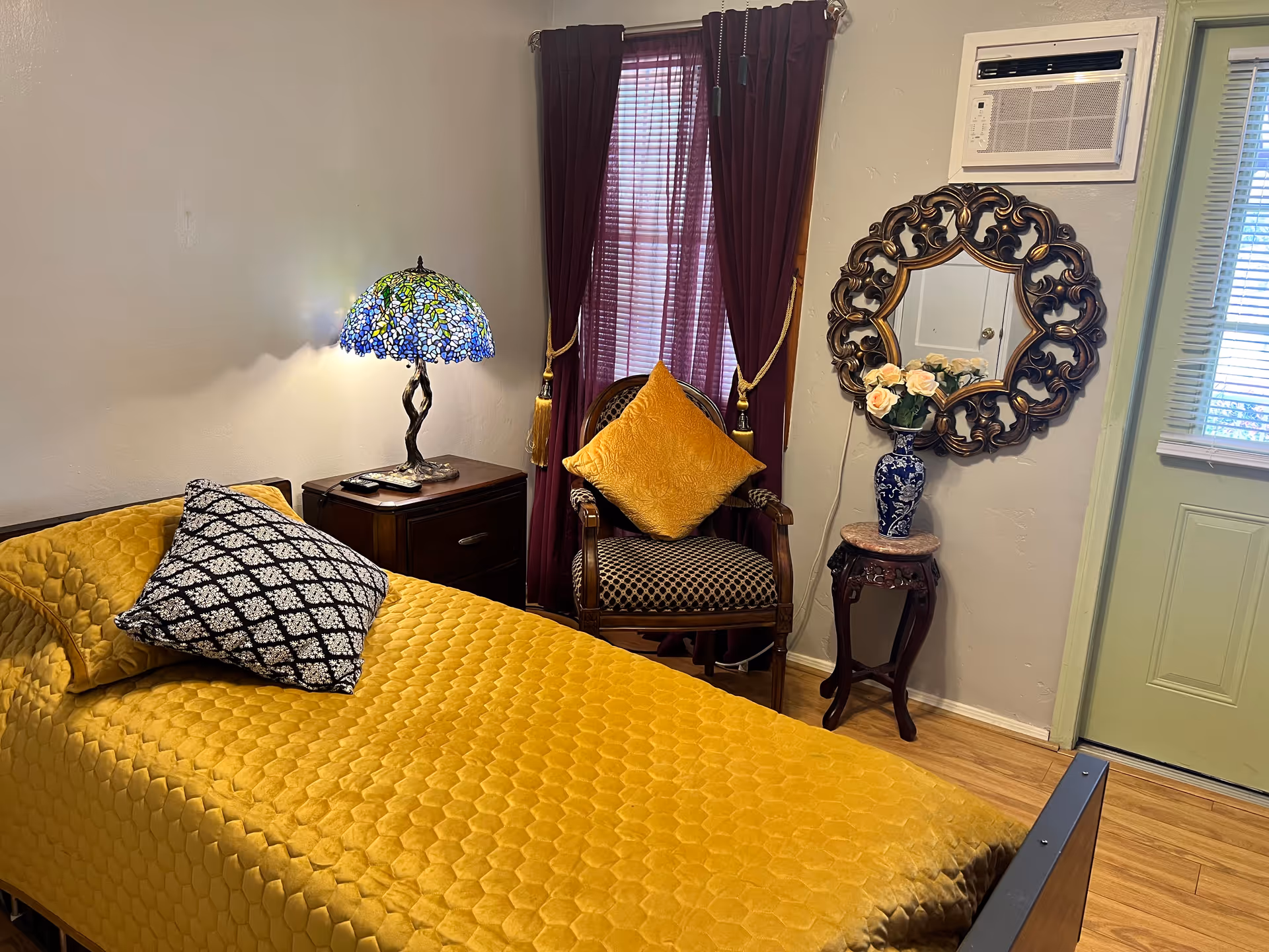 A cozy bedroom with a single bed covered in a mustard yellow quilt and a black and white patterned pillow. Next to the bed is a wooden nightstand with a decorative lamp featuring a blue and green stained glass shade. A wooden chair with a mustard yellow cushion is placed in front of a window with dark purple curtains. On the wall, there is an ornate round mirror above a small wooden table holding a blue vase with light pink flowers. An air conditioning unit is installed above the mirror, and a door with a window is visible on the right side.