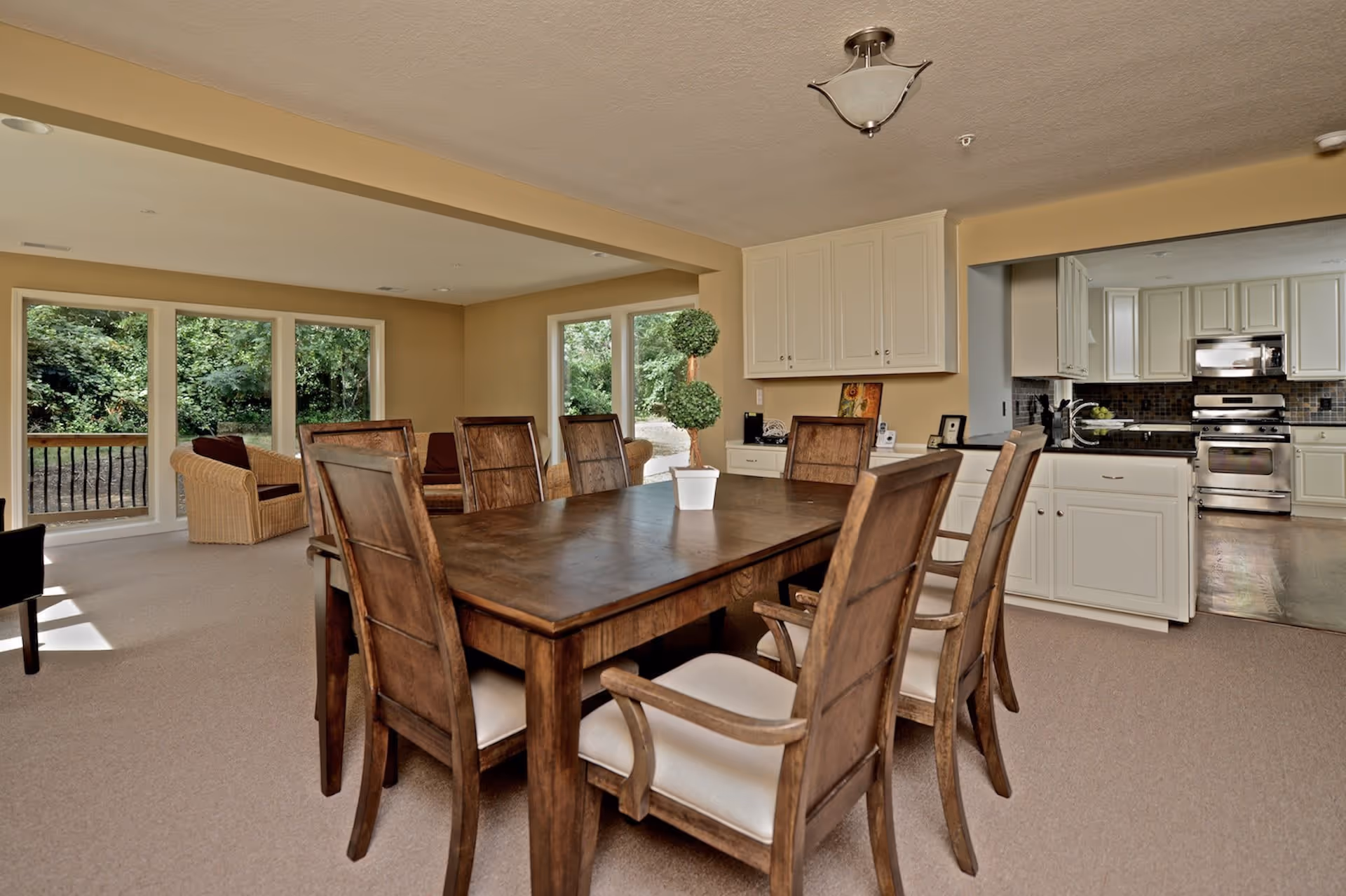 Dining room with a large wooden table and chairs opening to a kitchen and a sitting area with large windows showing greenery outside.