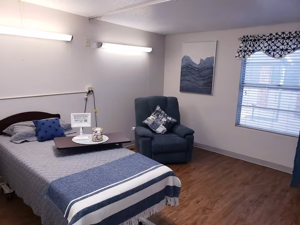 A nursing facility bedroom with a single bed covered in gray and blue bedding, a small overbed table with a decorative candle and a sign, a blue armchair with a patterned pillow, a window with blinds and a valance, and a framed abstract artwork on the wall.