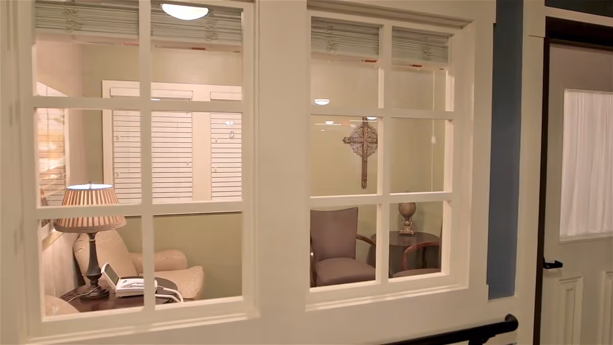 View through a multi-pane interior door into a small sitting room with upholstered chairs, a table lamp, a telephone, and a decorative wall cross.