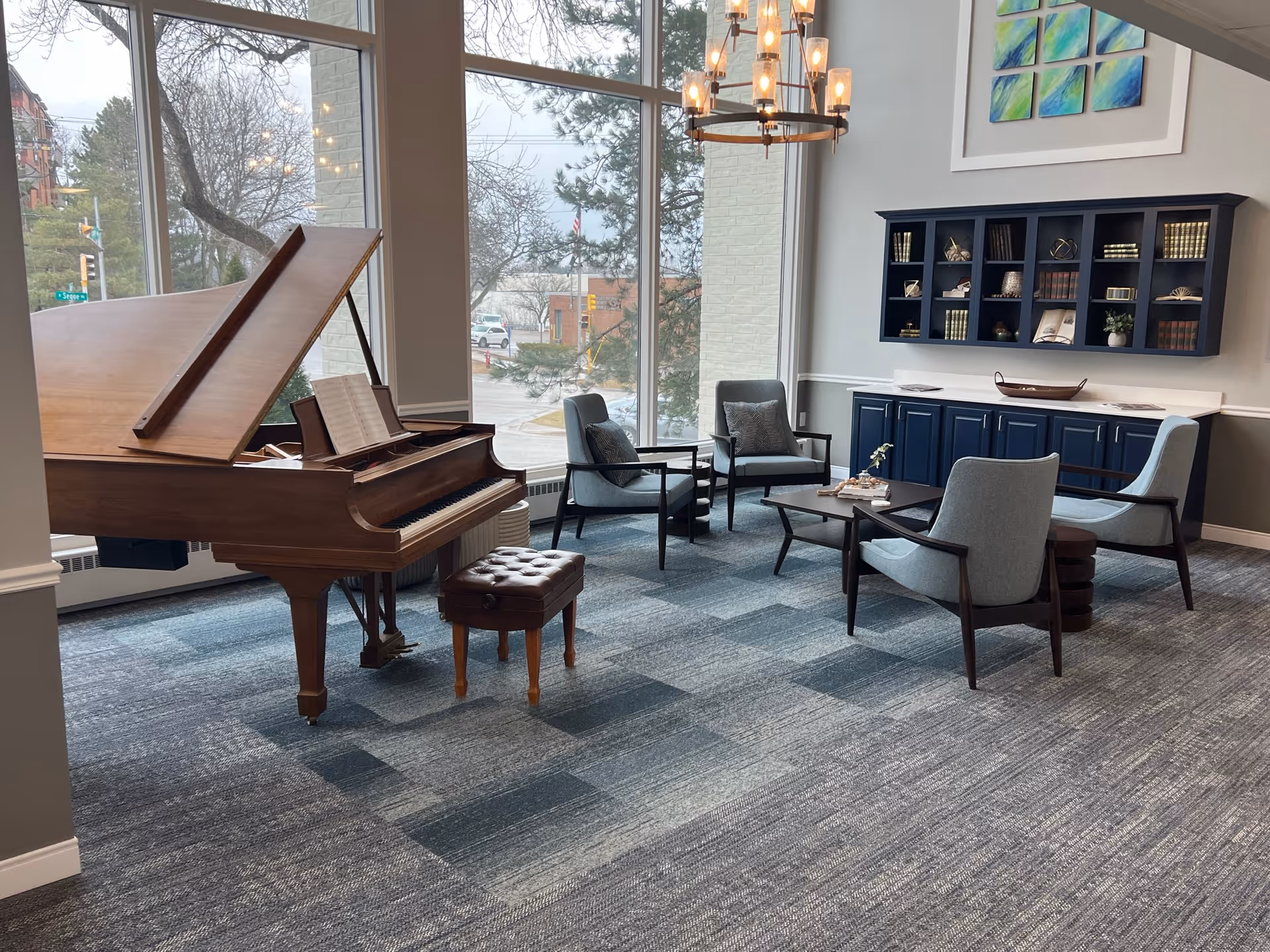 A bright and spacious lounge area with large floor-to-ceiling windows showing an outdoor street view. Inside, there is a wooden grand piano with an open lid and a cushioned piano bench. Nearby, four modern armchairs surround a low coffee table with decorative items. A blue cabinet with shelves filled with books and decor is mounted on the wall. A chandelier with multiple lights hangs from the ceiling.